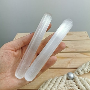 May include: Two white selenite wands, also known as selenite sticks, are held in a hand. The wands are smooth and have a striated texture.