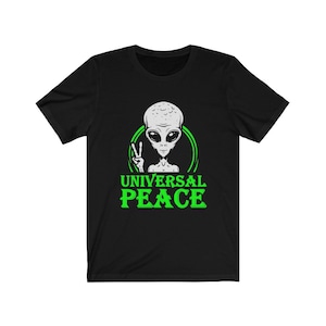 Puede incluir: Camiseta negra con un círculo verde y un extraterrestre blanco en su interior. El extraterrestre está haciendo un signo de paz con su mano. El texto "UNIVERSAL PEACE" está escrito en verde debajo del extraterrestre.