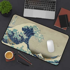 Puede incluir: Un ratón de ordenador blanco sobre una alfombrilla de ratón con un diseño de ola japonesa azul y blanca. La alfombrilla de ratón está sobre una superficie negra con un portátil, lápices y una libreta.