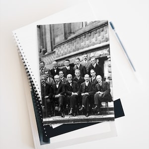May include: A black and white photograph of a group of men, likely scientists, gathered for the 5th Solvay International Conference on Electrons and Photons, held in Brussels, Belgium in October 1927. The men are seated in rows, with some standing behind them. The photo is taken outdoors, with a brick wall in the background.