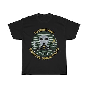 Könnte beinhalten: Schwarzes T-Shirt mit einem grünen und goldenen kreisförmigen Grafik. Die Grafik zeigt einen Außerirdischen, der ein Lenkrad hält, mit dem Text "To Serve Man" oben und "Gustatus Similis Pullus 509" unten.