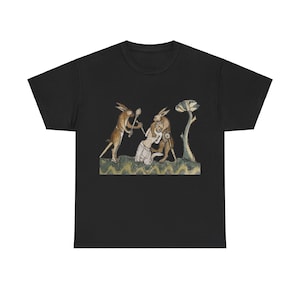 Puede incluir: Una camiseta negra con una ilustración de estilo medieval de tres conejos. Un conejo sostiene un palo y ataca a una persona, mientras que otro conejo ataca a la persona por detrás. La ilustración está sobre un fondo verde.