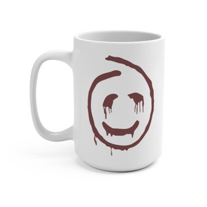Puede incluir: Taza de cerámica blanca con un diseño de cara sonriente marrón. El diseño presenta un contorno circular con detalles goteantes. La taza tiene un asa grande, ideal para bebidas calientes.