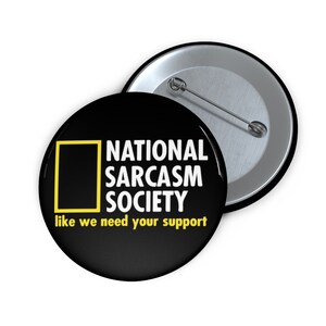 May include: Black button with white text that reads "National Sarcasm Society like we need your support". A yellow square with a black interior is in the upper left corner of the button.