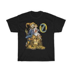 May include: Black t-shirt featuring a vintage illustration of Dorothy, the Scarecrow, and the Cowardly Lion from The Wizard of Oz. The illustration shows the characters riding on the lion, with Dorothy holding a basket and the Scarecrow holding a broom. The letter "O" from the word "Oz" is in the background.