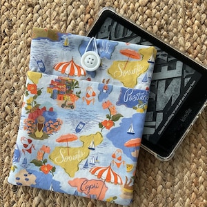 May include: A blue and white fabric Kindle cover with a button closure. The cover features a map design with orange umbrellas, sailboats, and the names of Italian coastal towns. A Kindle device is partially visible.