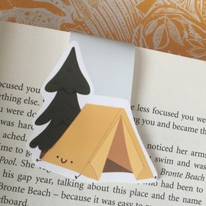 May include: A sticker featuring a cartoon illustration of a tent and a pine tree. The tent is brown and yellow with a smiling face. The pine tree is black with a white smiling face. The sticker is on a book with text about Bronte Beach.