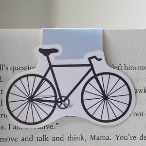 May include: A black and white bicycle bookmark with a light blue background. The bookmark is shaped like a bicycle with two wheels, a handlebar, and a seat.