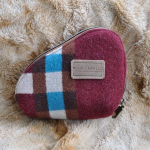 Puede incluir: Una bolsa con cremallera burdeos y a cuadros con una etiqueta de cuero que dice "WILD CABALLO". La bolsa tiene un estampado de cuadros en blanco, azul y marrón. El fondo es una superficie borrosa de color marrón claro.