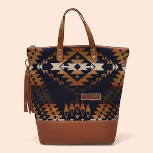 Peut inclure: Un sac fourre-tout avec un motif géométrique bleu marine, beige et blanc. Le sac a des poignées en cuir marron et un fond en cuir. Une gland en cuir marron est suspendu sur le côté. Le sac a une fermeture éclair.
