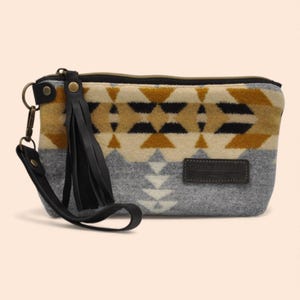 May include: A rectangular clutch with a geometric pattern in gray, tan, and black. It has a black leather tassel and wrist strap. A small, rectangular leather patch is sewn on the front.
