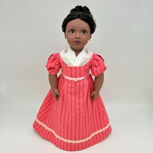 May include: A doll with dark brown hair styled in an updo, wearing a coral-colored dress with a repeating dark pattern, short puff sleeves, a white shawl collar, a thin pink ribbon waistband with a small center embellishment, and cream lace trim.