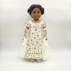 May include: A doll with dark brown hair and green eyes wearing a cream-coloured dress with a small purple floral pattern. The dress features a purple ribbon at the waist, short puffed sleeves with purple bows, and two ruffled tiers at the hem.