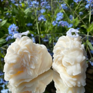 May include: Two white lion head candles with a detailed design. The candles are lying on a reflective surface with a blurred background of blue flowers.