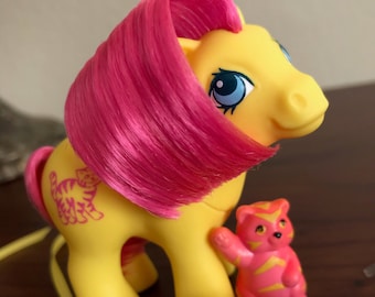 May include: A yellow pony with pink hair and a tiger design on its side. The pony is holding a small pink and orange tiger toy. There are also two cat-shaped rings and a small cake with a strawberry on top.