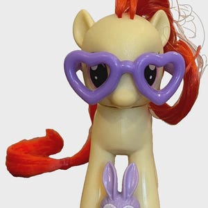 May include: A yellow toy pony figure with orange hair and purple heart-shaped glasses. A small purple bunny figure is in front of the pony. The pony has a long orange tail.