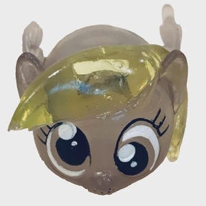 May include: A close-up of a cartoon character's head, with a light brown face and large eyes. The character has a yellow, translucent hair piece and small, clear wings. The eyes are outlined in black with white highlights.
