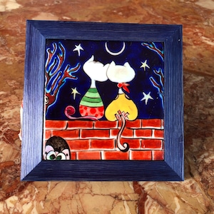 May include: A framed ceramic tile featuring two stylized white cats gazing at each other under a crescent moon and stars. The cats are perched on a red brick wall against a dark blue night sky. The frame is a dark blue.