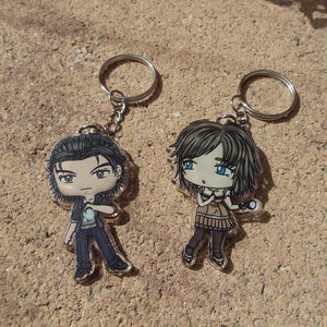 Pode incluir: Dois chaveiros com personagens de desenhos animados, um com um personagem masculino com cabelo preto e uma jaqueta preta, e o outro com um personagem feminino com cabelo castanho e um vestido marrom. Ambos os personagens têm olhos grandes e estão segurando um microfone.