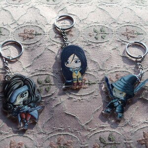 May include: Three keychains featuring anime characters. The first keychain features a girl with black hair and a blue dress. The second keychain features a girl with brown hair and a white dress. The third keychain features a character wearing a black hat and holding a sword.