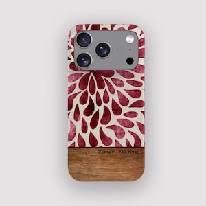 Può includere: Custodia per telefono con un design floreale. La parte superiore presenta uno sfondo color crema con un motivo di gocce bordeaux. La sezione inferiore ha un aspetto venato del legno. Il testo "Your Name" è stampato sulla venatura del legno.