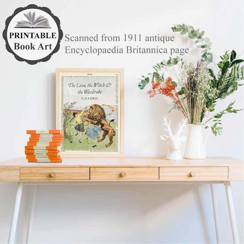 May include: A framed print of "The Lion, the Witch & the Wardrobe" on a vintage book page, with a stack of orange books. The print is on a light wood desk with a white vase of flowers and a deer head figurine. Text on the print reads "PRINTABLE Book Art" and "Scanned from 1911 antique Encyclopaedia Britannica page."