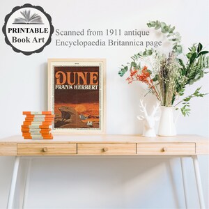 K&ouml;nnte beinhalten: Ein gerahmter Druck des Buchcovers f&uuml;r "Dune" von Frank Herbert. Das Cover zeigt eine W&uuml;stenlandschaft mit einem Sandwurm im Hintergrund. Der Druck befindet sich auf einem Holztisch mit einem Stapel B&uuml;cher und einer Vase mit Blumen.
