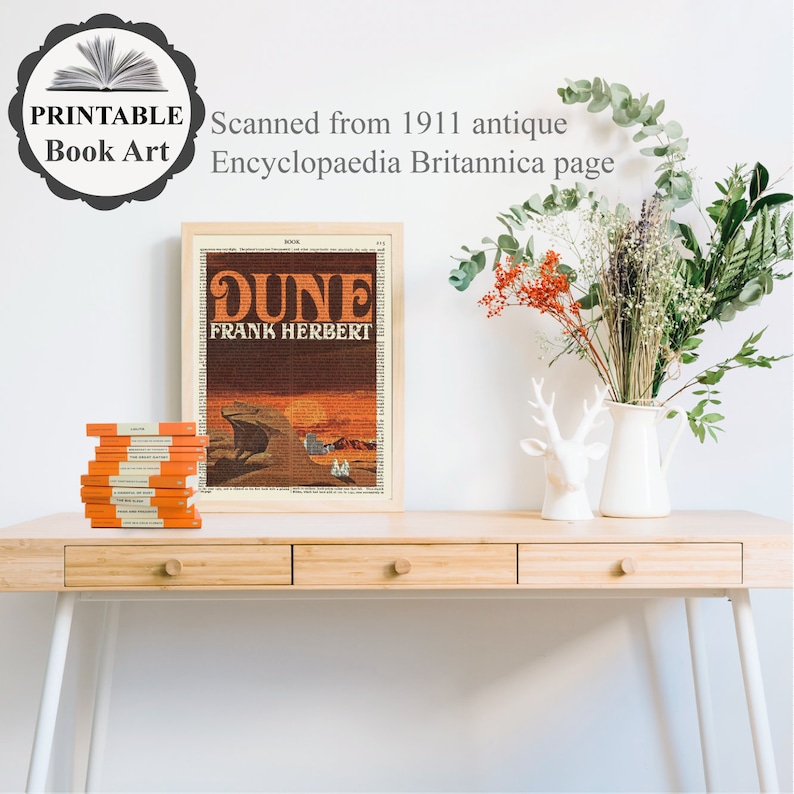 K&ouml;nnte beinhalten: Ein gerahmter Druck des Buchcovers f&uuml;r "Dune" von Frank Herbert. Das Cover zeigt eine W&uuml;stenlandschaft mit einem Sandwurm im Hintergrund. Der Druck befindet sich auf einem Holztisch mit einem Stapel B&uuml;cher und einer Vase mit Blumen.