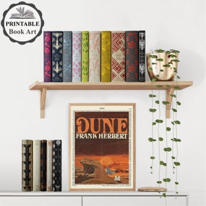 K&ouml;nnte beinhalten: Ein gerahmter Druck eines Buchcovers f&uuml;r "Dune" von Frank Herbert. Das Cover ist eine Collage aus Text und einer W&uuml;stenszene mit einem Sandwurm. Der Druck wird in einem Regal mit einer Sammlung bunter B&uuml;cher und einer H&auml;ngepflanze ausgestellt.