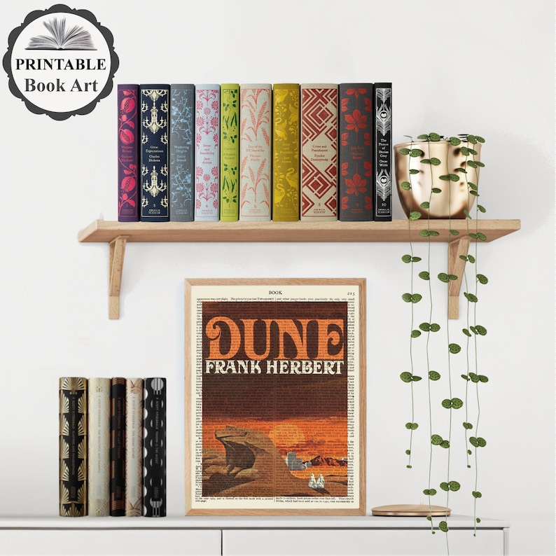K&ouml;nnte beinhalten: Ein gerahmter Druck eines Buchcovers f&uuml;r "Dune" von Frank Herbert. Das Cover ist eine Collage aus Text und einer W&uuml;stenszene mit einem Sandwurm. Der Druck wird in einem Regal mit einer Sammlung bunter B&uuml;cher und einer H&auml;ngepflanze ausgestellt.