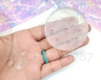 May include: A clear, circular mold with multiple small holes, labeled "CRAFTY 787". Three small, clear, circular objects are next to the mold. The background is white and iridescent.