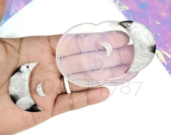 May include: A clear silicone mold with two crescent moon shapes. The mold is being held in a hand, and there are two finished resin pieces next to it. The resin pieces are silver and black.