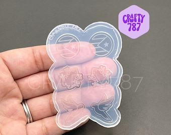 May include: Clear silicone mold with multiple cavities. The mold features designs of a flag with a star, frogs, and doves. The text "Crafty 787" is visible in a purple hexagon. The mold is held in a hand.