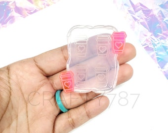 May include: Clear silicone mold with six heart-shaped coffee cup designs. The mold is held in a hand with a turquoise ring. Three pink coffee cups are shown outside of the mold.