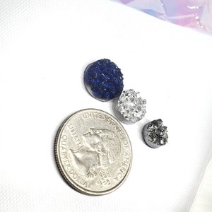 May include: Three druzy-style cabochons in navy blue, silver, and charcoal gray, next to a quarter. The cabochons have a textured, crystalline surface. The quarter is included for size reference.