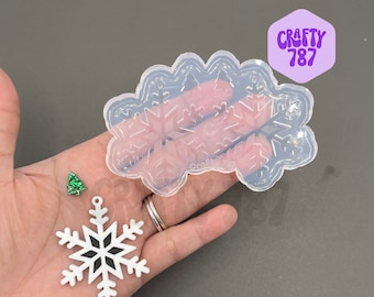 Op de afbeelding: Een transparante siliconen mal met een ontwerp van meerdere sneeuwvlokken. Naast de mal is een witte sneeuwvlok charme te zien. De mal is gelabeld met "CRAFTY 787".