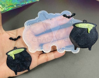 Könnte beinhalten: Eine transparente Silikonform mit einem Kessel- und Fledermaus-Design, gehalten in einer Hand. Zwei schwarze Kessel mit grüner Flüssigkeit und zwei schwarze Fledermäuse befinden sich neben der Form. Das Wort "Crafty78" ist sichtbar.