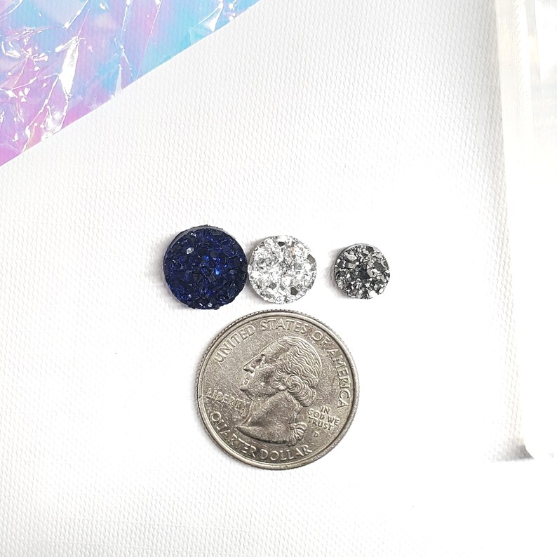 May include: Three round, sparkly cabochons in navy blue, silver, and gray, next to a quarter. The cabochons vary in size, with the navy blue one being the largest. The quarter is included for size reference.