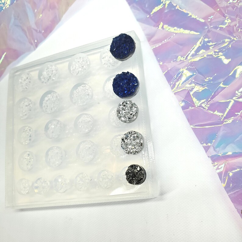 May include: A clear plastic tray filled with small, round, clear objects. Four pairs of colorful, sparkly stud earrings are displayed on the right side of the tray. The earrings are dark blue, silver, and black.