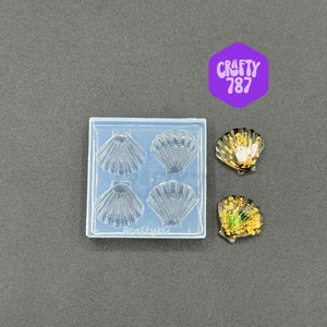 Può includere: Uno stampo in silicone trasparente con quattro impronte di conchiglie e due pezzi di resina a forma di conchiglia finiti. Le conchiglie in resina sono dorate, verdi e nere. L'immagine include un esagono viola con il testo "CRAFTY 787".