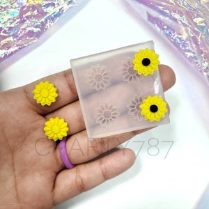 Puede incluir: Un molde de silicona transparente con cuatro formas de girasol. Se muestran tres pendientes de girasol amarillos con centros negros junto al molde.