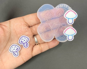 May include: A clear silicone mold with six mushroom shapes. The mold is on a hand holding two white resin mushrooms with purple outlines and a crescent moon and stars design.