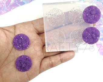 May include: A clear silicone mold with four round cavities for making resin crafts. Two purple resin pieces with a sparkly, crystal-like texture are shown next to the mold.
