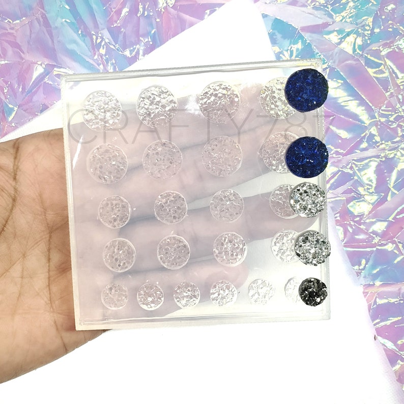 May include: A clear plastic mold filled with various sizes of round, textured cabochons. Some cabochons are clear, while others are dark blue, silver, and black. The word "CRAFTY" is partially visible on the mold.