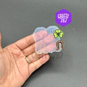May include: Clear silicone mold with four horseshoe shapes and two four-leaf clover shapes. The mold is held in a hand.