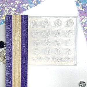 May include: A clear, rectangular mold with 20 spherical cavities of varying sizes. A wooden ruler with inch markings is positioned next to the mold. The mold is likely for crafting decorative elements.