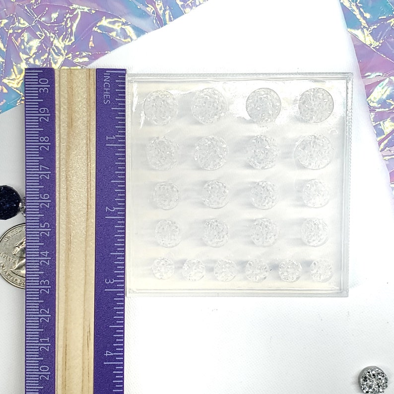 May include: A clear, rectangular mold with 20 spherical cavities of varying sizes. A wooden ruler with inch markings is positioned next to the mold. The mold is likely for crafting decorative elements.