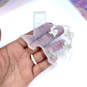 May include: Clear silicone mold shaped like the state of Texas with smaller Texas shapes inside. The mold is on a hand with a white ring.