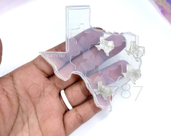 May include: Clear silicone mold shaped like the state of Texas with smaller Texas shapes inside. The mold is on a hand with a white ring.