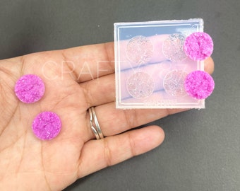 May include: A clear silicone mold with four circular cavities, each with a detailed design. Three bright pink, textured, circular resin pieces are next to the mold. The image is on a dark gray surface.